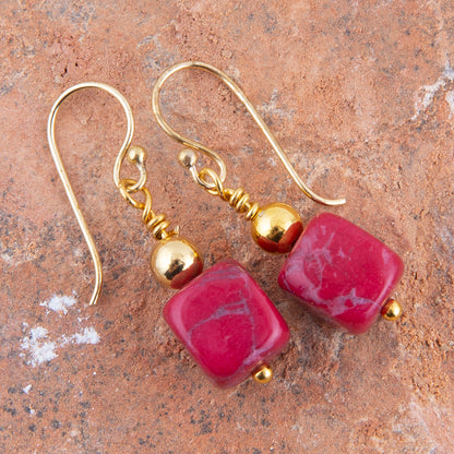 A pair of gold-tone dangle earrings with square pink gemstone beads, displayed on a textured terracotta surface.