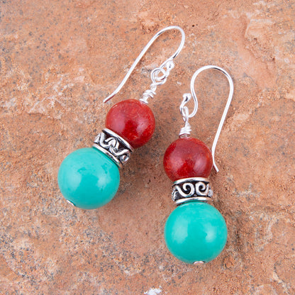 A pair of dangle earrings with turquoise-green and red beads separated by silver filigree spacers, displayed on a textured brown stone surface.
