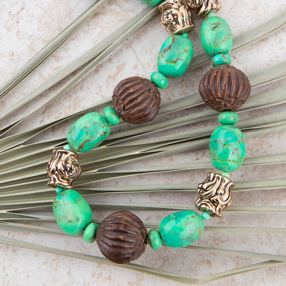 A beaded necklace featuring turquoise-green marbled beads, dark brown ribbed beads, and antique gold-toned lion head spacers, laid flat on a light stone surface with dried palm leaves.