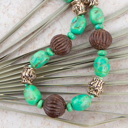 A beaded necklace featuring turquoise-green marbled beads, dark brown ribbed beads, and antique gold-toned lion head spacers, laid flat on a light stone surface with dried palm leaves.