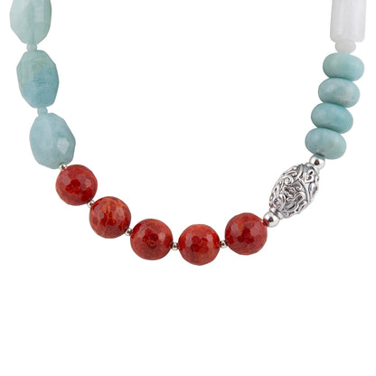 A beaded necklace featuring faceted light blue beads, five round red beads, a textured silver spacer, and a white cylindrical bead on a plain white background.
