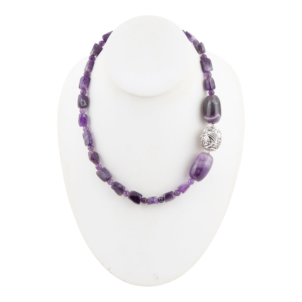 A beaded necklace made of purple amethyst stones and a textured silver accent bead, displayed on a white mannequin bust against a white background.