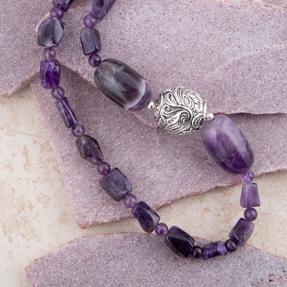 A beaded necklace featuring large oval and rectangular purple amethyst beads, small round purple beads, and a prominent ornate silver filigree bead, displayed on textured stone tiles.