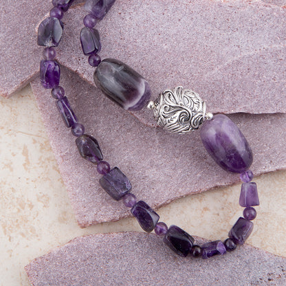 A beaded necklace featuring large oval and rectangular purple amethyst beads, small round purple beads, and a prominent ornate silver filigree bead, displayed on textured stone tiles.