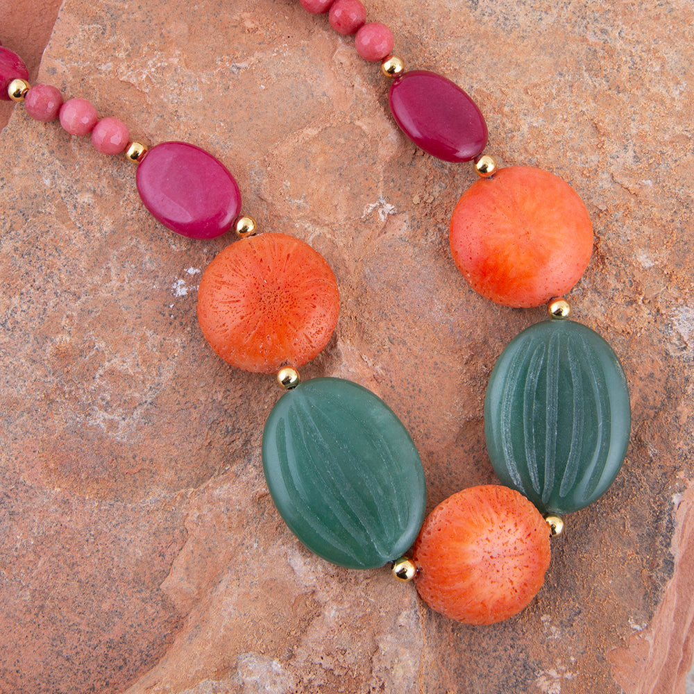 A beaded necklace with large oval and round beads in pink, orange, and green, separated by small gold spacer beads, displayed on a textured brown stone surface.