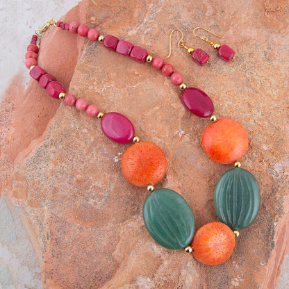 A colorful beaded necklace with matching drop earrings displayed on a textured brown rock surface. The necklace features pink and coral-orange round beads, dark green ribbed oval beads, and shiny magenta oval beads separated by small gold spacer beads.