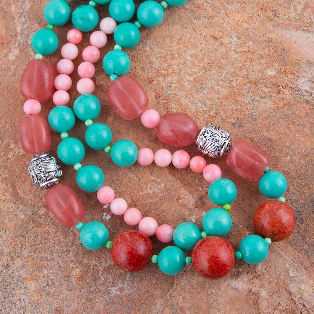 A beaded necklace with alternating turquoise, coral red, and light pink beads, accented by silver-toned textured spacers, displayed on a textured brown stone surface.