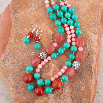 A beaded necklace and matching drop earrings laid flat on a textured brown rock. The jewelry features turquoise, coral pink, and reddish-orange beads with silver-toned spacer details.