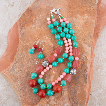 A beaded necklace and matching drop earrings laid flat on a textured brown rock. The jewelry features turquoise, coral pink, and reddish-orange beads with silver-toned spacer details.