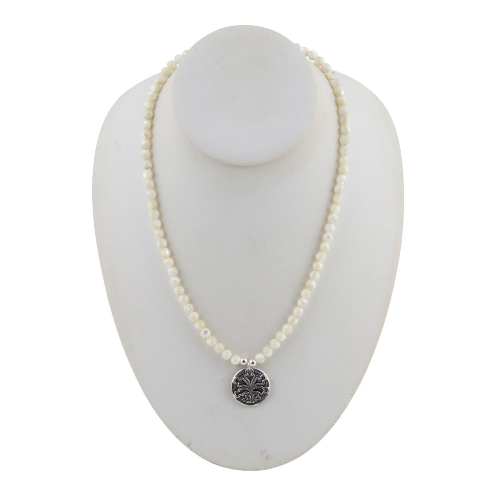 A white pearl beaded necklace with a silver-toned circular pendant featuring an engraved floral or sunburst pattern, displayed on a white mannequin bust against a white background.