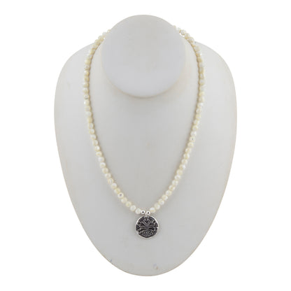 A white pearl beaded necklace with a silver-toned circular pendant featuring an engraved floral or sunburst pattern, displayed on a white mannequin bust against a white background.