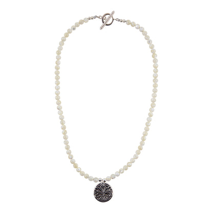 A beaded necklace featuring small, round off-white beads and a silver-toned toggle clasp, with a circular silver pendant engraved with a floral pattern, shown against a plain white background.