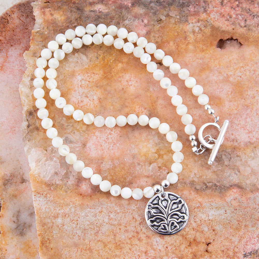 A beaded necklace with round off-white beads, a silver toggle clasp, and a round silver pendant featuring an engraved floral or tree-like motif, displayed on a textured orange-brown stone surface.