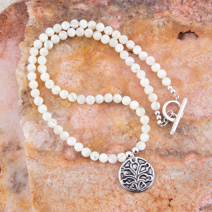 A beaded necklace with round off-white beads, a silver toggle clasp, and a round silver pendant featuring an engraved floral or tree-like motif, displayed on a textured orange-brown stone surface.