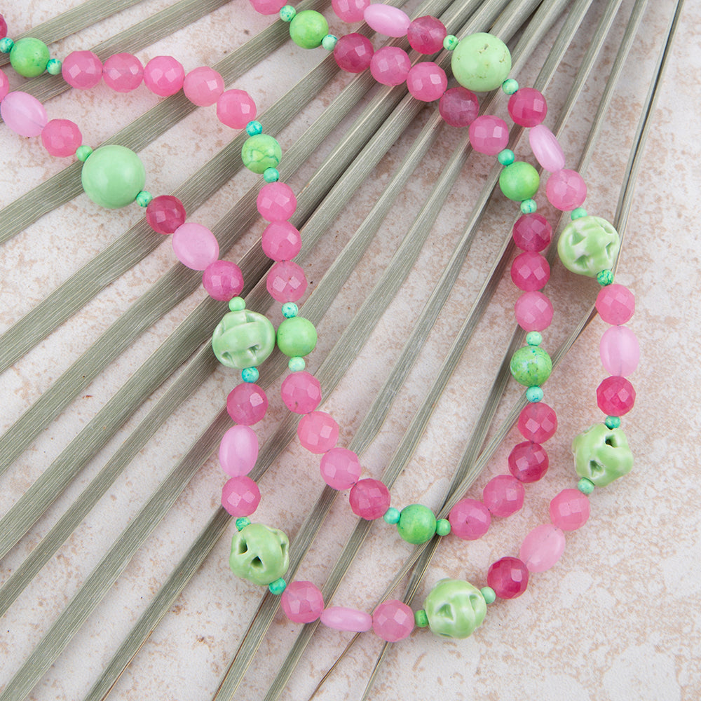 A beaded necklace with pink and light green beads, including faceted round beads, smooth oval beads, and carved bell-shaped beads, displayed on a fan of green palm leaves over a light textured background.