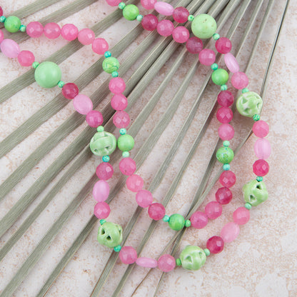 A beaded necklace with pink and light green beads, including faceted round beads, smooth oval beads, and carved bell-shaped beads, displayed on a fan of green palm leaves over a light textured background.