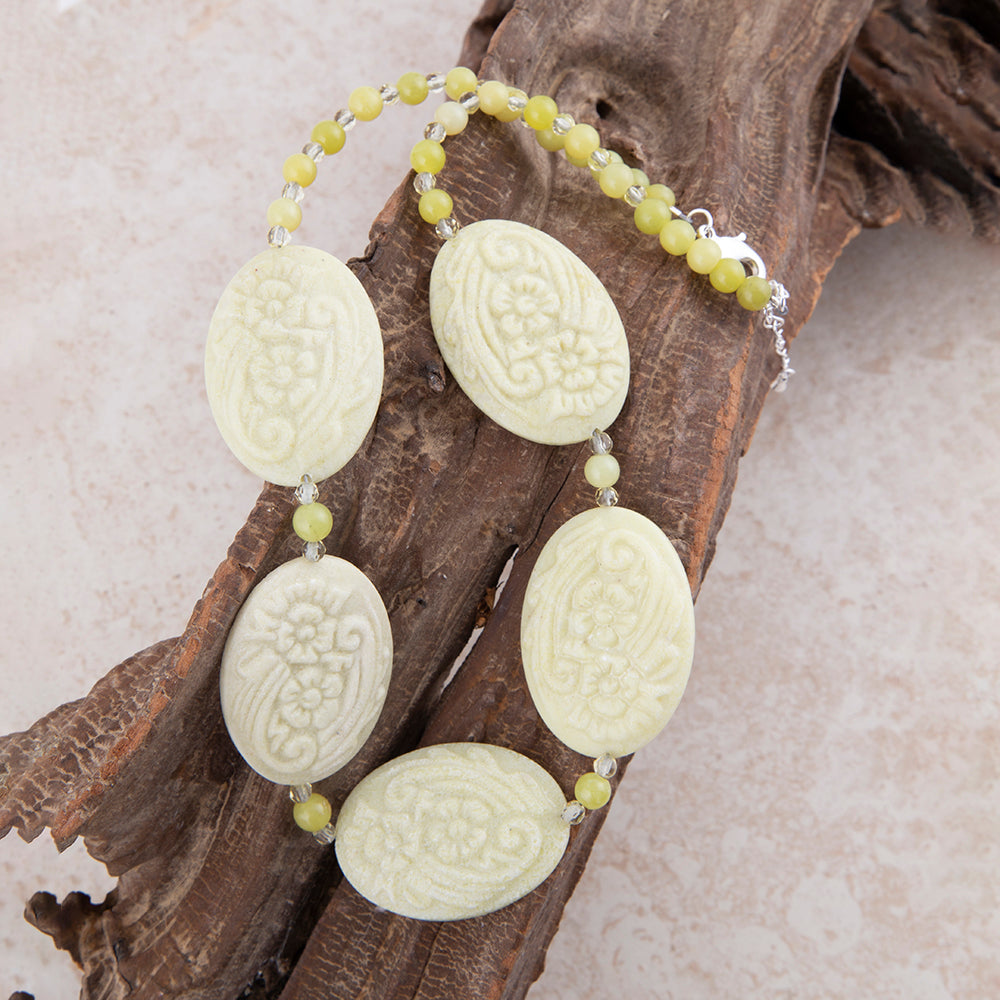 A beaded necklace with five carved oval pale yellow-green stone pendants and small round beads, displayed on a piece of driftwood against a light beige background.