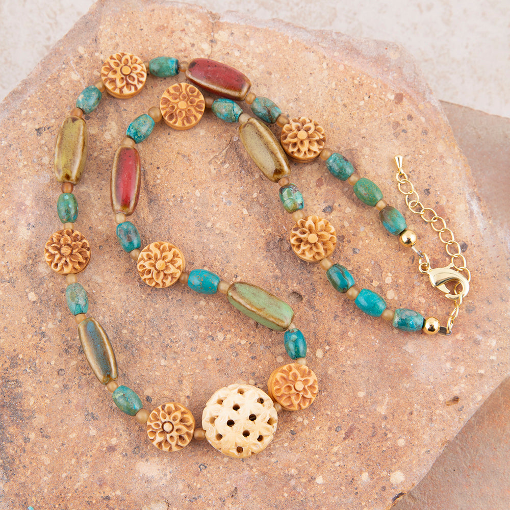 A beaded necklace with turquoise-blue beads, carved gold-toned floral beads, and a red rectangular bead, displayed on a textured tan stone surface.