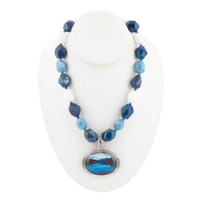 A beaded necklace displayed on a white mannequin bust, featuring faceted beads in navy, light blue, and white, with a silver-tone oval pendant depicting a blue and black landscape.