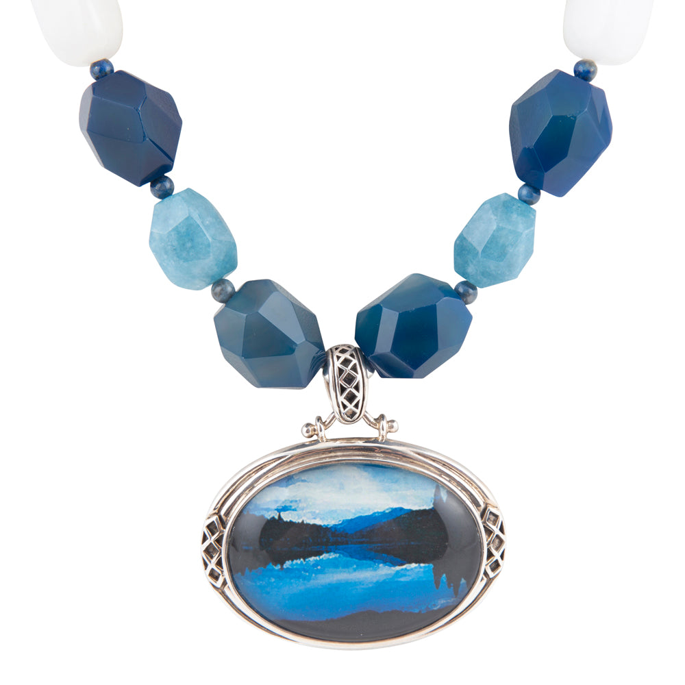 A beaded necklace with faceted blue and light blue beads, white end beads, and a large oval pendant featuring a blue and black landscape painting in a silver-toned frame, shown on a white background.