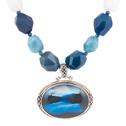A beaded necklace with faceted blue and light blue beads, white end beads, and a large oval pendant featuring a blue and black landscape painting in a silver-toned frame, shown on a white background.