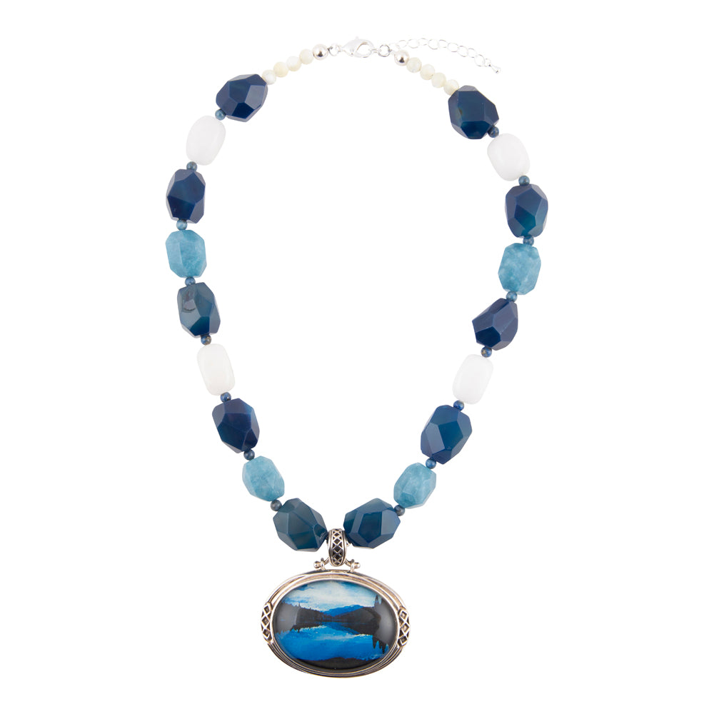 A beaded necklace featuring faceted blue and light blue beads, white translucent beads, and a large oval pendant with a blue and black landscape scene, displayed on a clean white background.