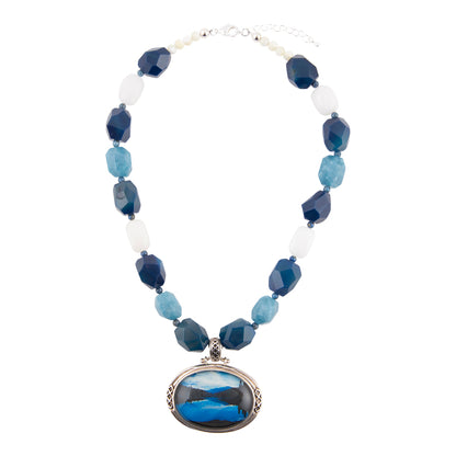 A beaded necklace featuring faceted blue and light blue beads, white translucent beads, and a large oval pendant with a blue and black landscape scene, displayed on a clean white background.