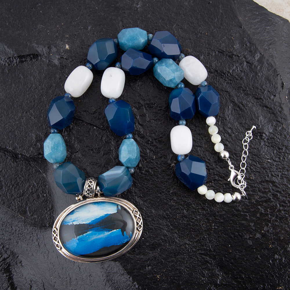 A beaded necklace with faceted blue and light blue beads, white beads, and a large oval pendant featuring a blue and white abstract design in a silver frame, displayed on a dark textured stone background.