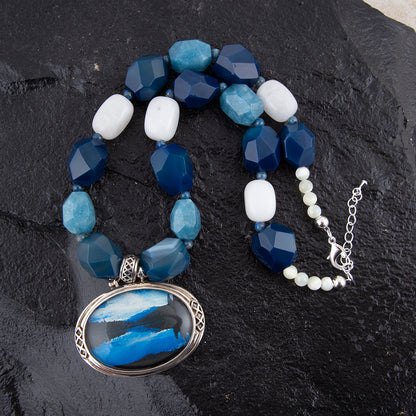 A beaded necklace with faceted blue and light blue beads, white beads, and a large oval pendant featuring a blue and white abstract design in a silver frame, displayed on a dark textured stone background.