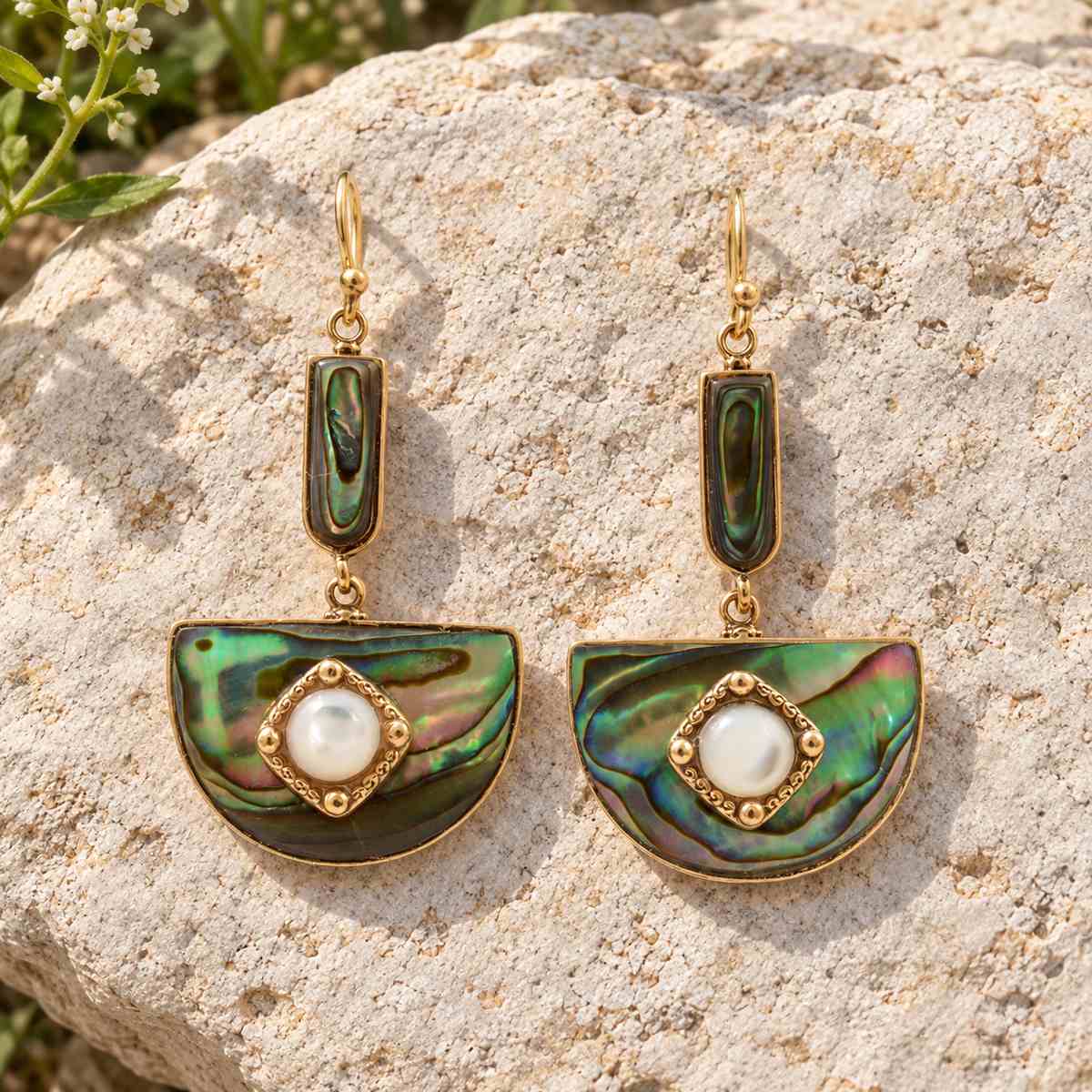 A pair of gold-tone dangle earrings with iridescent green and blue abalone shell semicircles and white pearl centers, displayed on a textured beige rock outdoors.