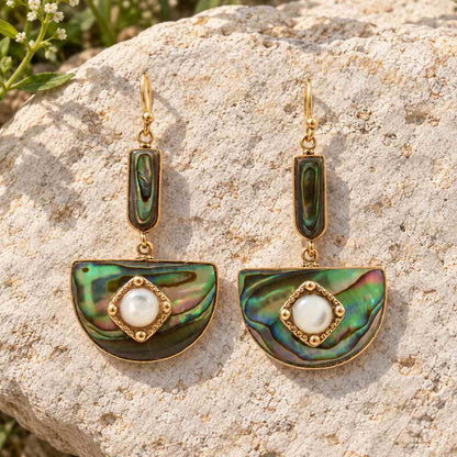 A pair of gold-tone dangle earrings with iridescent green and blue abalone shell semicircles and white pearl centers, displayed on a textured beige rock outdoors.