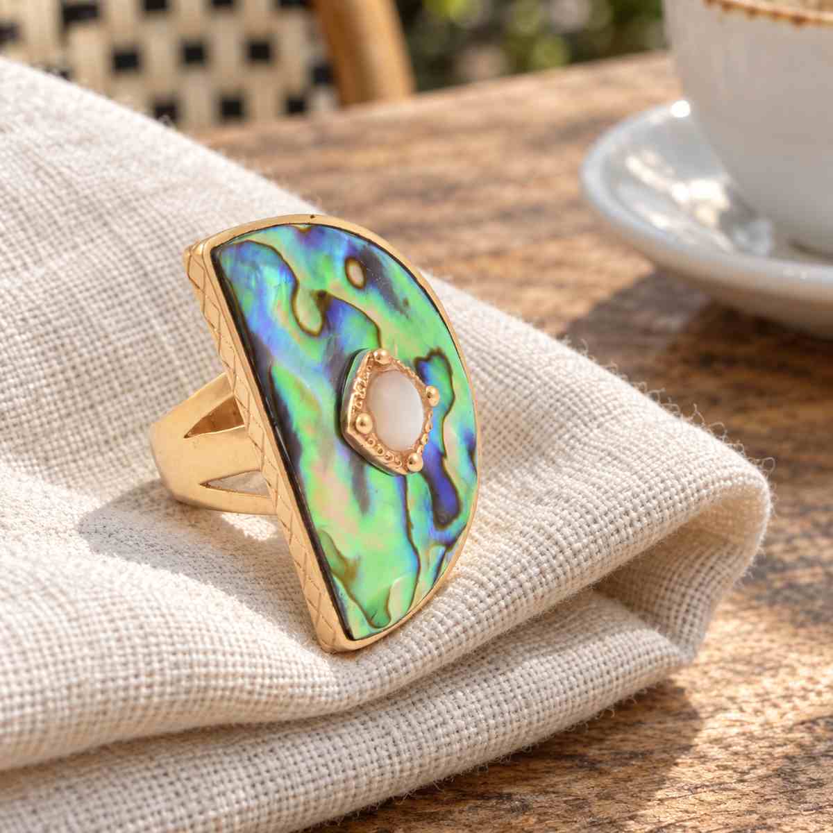 A gold ring with a large, irregular-shaped green and blue iridescent abalone shell inlay and a central white stone, resting on a beige linen cloth on a wooden table with a blurred coffee cup in the background.