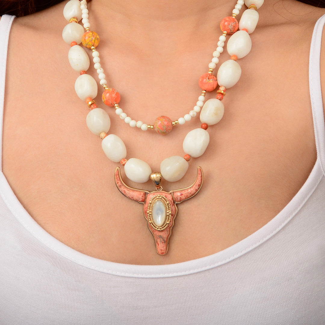 Necklace with a large pink bull head pendant and white and orange beads on a person wearing a white top.