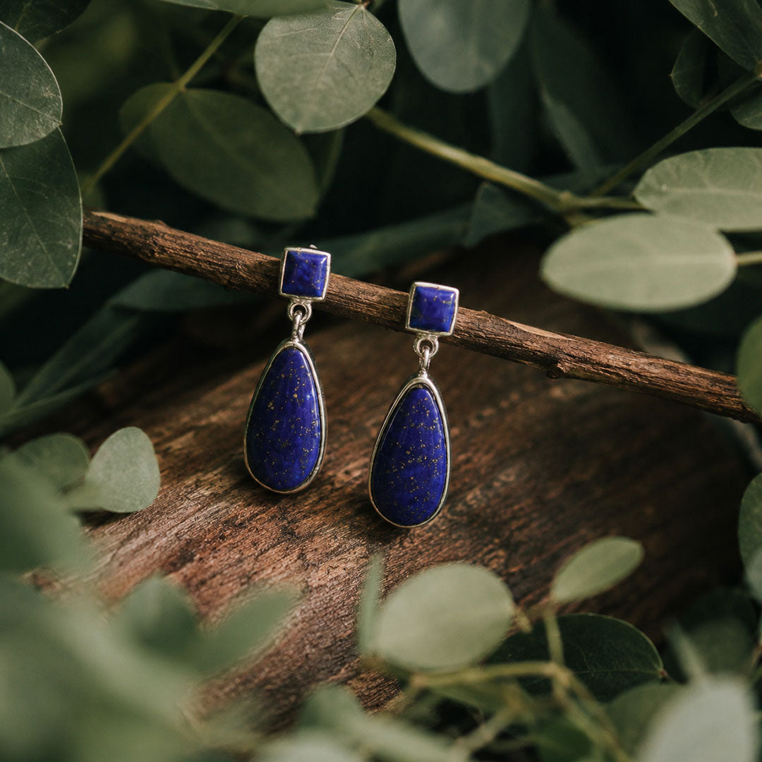 A pair of silver drop earrings with deep blue lapis lazuli stones, displayed on a rustic wooden branch surrounded by green leaves.
