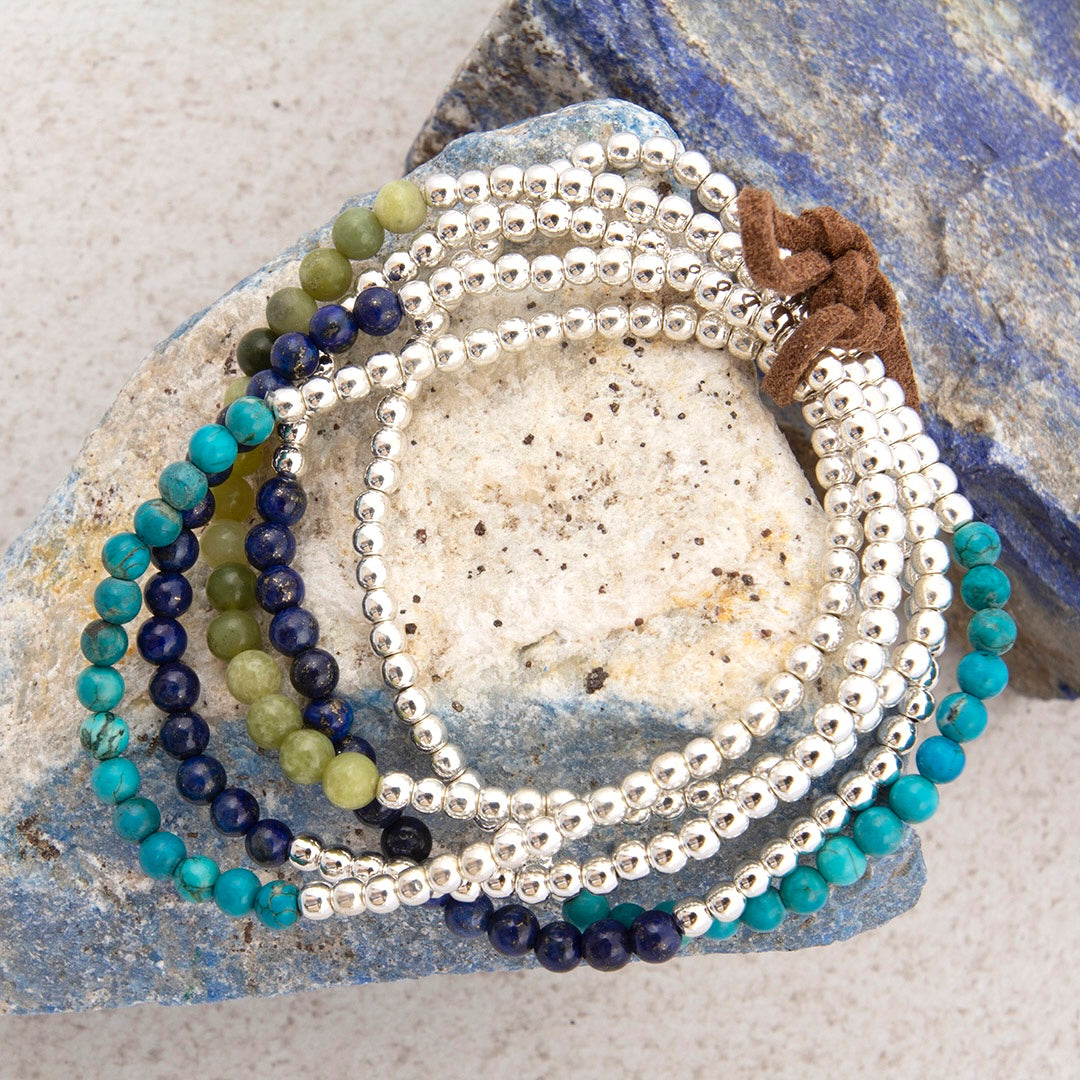 A set of beaded bracelets arranged on a stone surface. The bracelets include silver-toned round beads, turquoise-blue beads, dark navy beads, olive-green beads, and a brown cord accent.