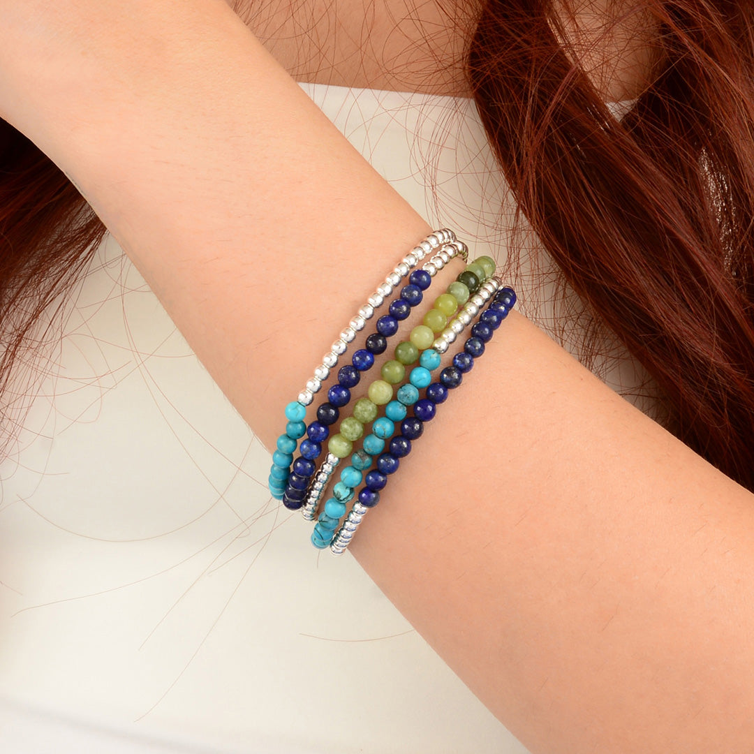 A close-up of a person's wrist wearing a stack of beaded stretch bracelets in shades of blue, green, turquoise, and silver.