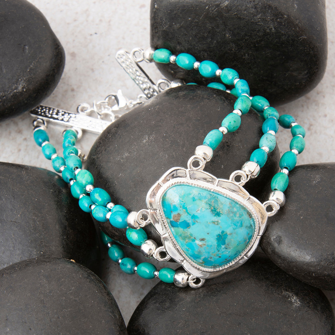 A beaded bracelet with turquoise-blue beads and a large triangular turquoise stone pendant in a silver frame, arranged on dark smooth rocks.