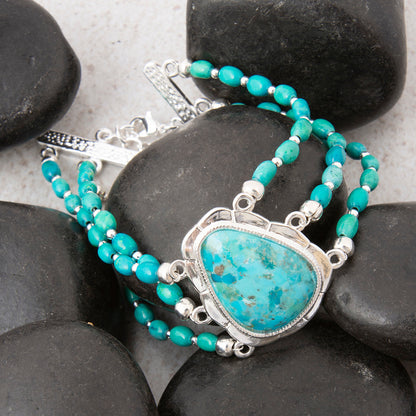 A beaded bracelet with turquoise-blue beads and a large triangular turquoise stone pendant in a silver frame, arranged on dark smooth rocks.
