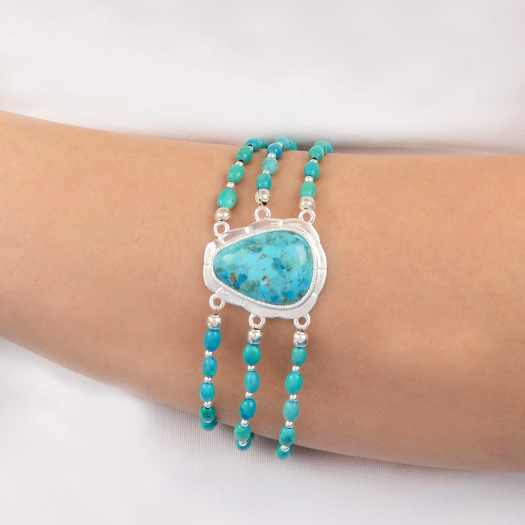 A close-up of a person's wrist wearing a multi-strand turquoise bracelet with a large central turquoise stone in a silver bezel setting and small silver beads.