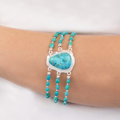 A close-up of a person's wrist wearing a multi-strand turquoise bracelet with a large central turquoise stone in a silver bezel setting and small silver beads.