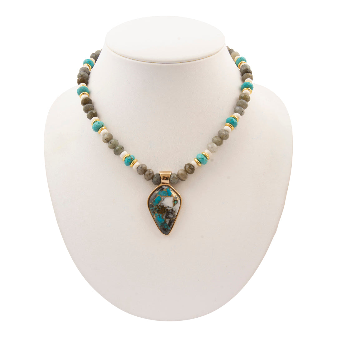 A beaded necklace displayed on a white jewelry bust. The necklace features a mix of gray and turquoise beads with small gold spacer beads, and a central teardrop-shaped pendant with a blue and black marbled pattern set in gold.