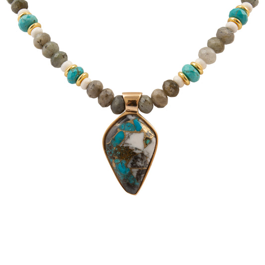 A beaded necklace featuring a central teardrop-shaped turquoise stone pendant in a gold bezel setting, flanked by gray stone beads, white beads, and small turquoise accent beads separated by gold spacers, on a plain white background.