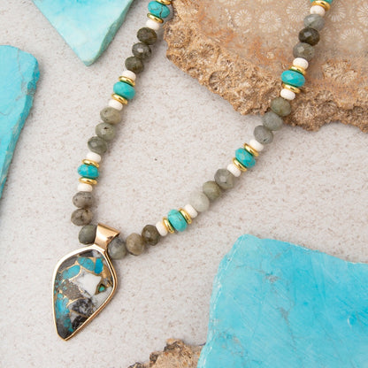 A beaded necklace with a teardrop-shaped pendant, featuring gray and turquoise beads, white accents, and gold-toned metal, displayed on a textured light gray surface with decorative turquoise and beige stones.
