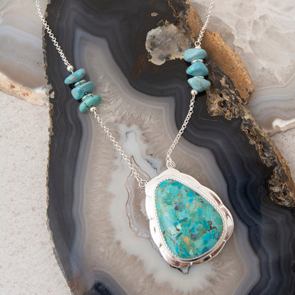 A silver necklace with a large turquoise-blue agate pendant and three small turquoise beads on the chain, displayed on a polished agate slice.