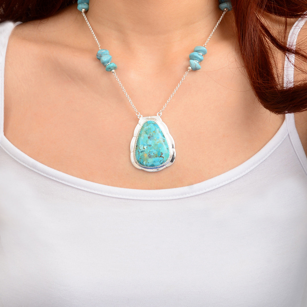 A close-up of a woman wearing a silver necklace with a large teardrop-shaped turquoise stone pendant and matching turquoise beads on the chain.