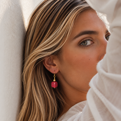 A close-up lifestyle shot of a woman with blonde-highlighted hair wearing a single drop earring. The earring features a small gold-toned bead and a dangling pink faceted stone, set against a soft white textured background.