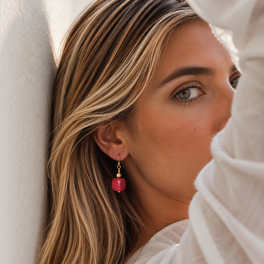 A close-up lifestyle shot of a woman with blonde-highlighted hair wearing a single drop earring. The earring features a small gold-toned bead and a dangling pink faceted stone, set against a soft white textured background.