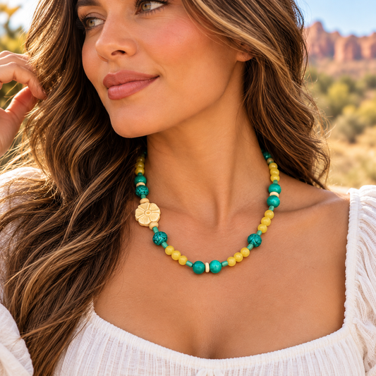 A woman wearing a colorful beaded necklace with yellow and green beads, turquoise accents, and a gold clover charm, photographed outdoors against a blurred desert landscape.