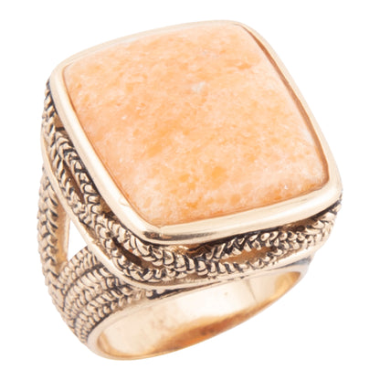 A square bezel-set ring featuring a textured, warm peach-orange stone, set in an ornate, braided gold-tone metal band with a polished gold inner band, shown against a plain white background.