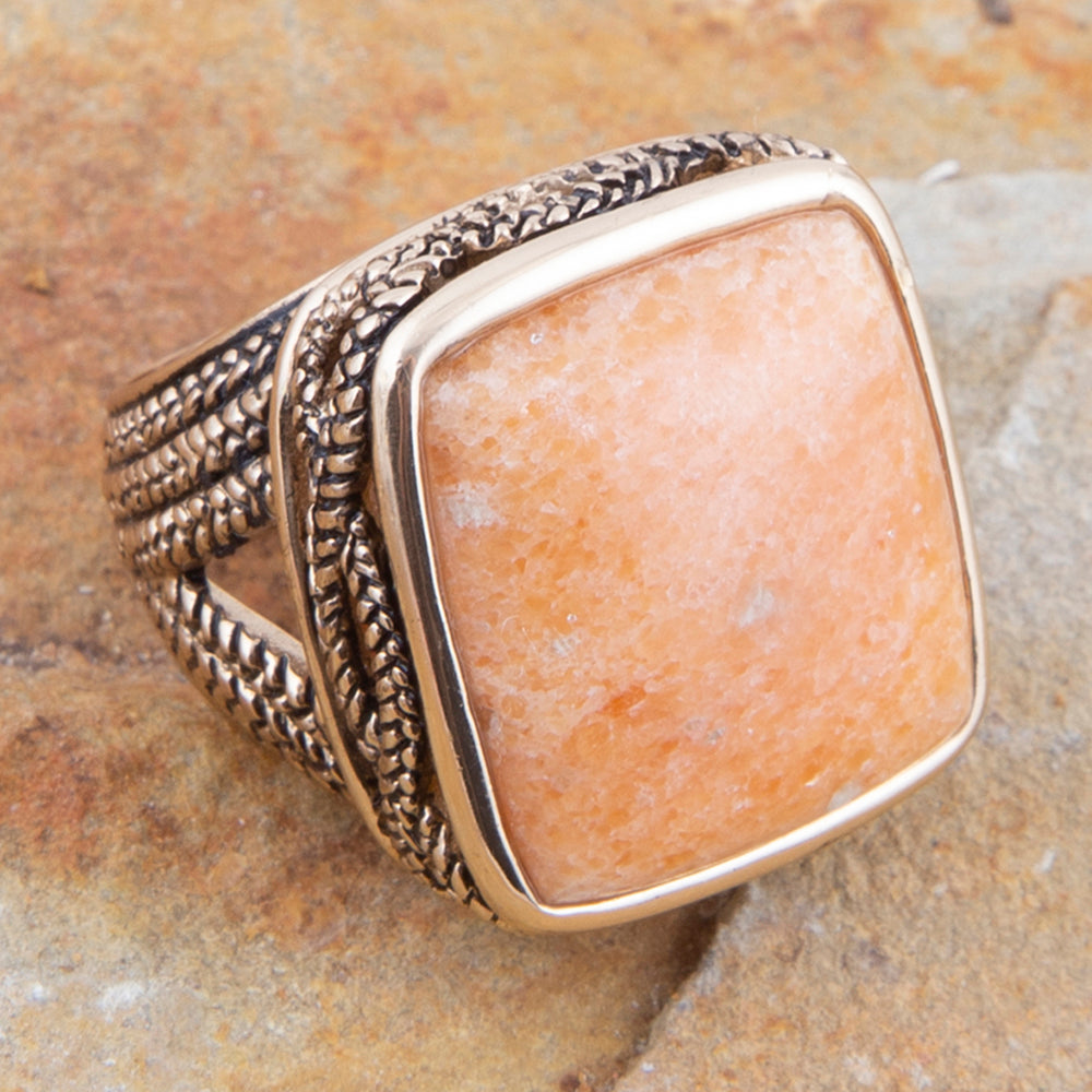 A square bezel-set ring featuring a warm peach-orange stone, set in a textured silver-toned metal band with a polished gold-toned bezel, resting on a textured stone surface.